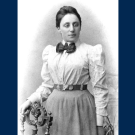 Black and white photo of a woman in early 20th century clothing with a white blouse, long skirt and bow at her throat. Her hand rests on the back of a chair. 